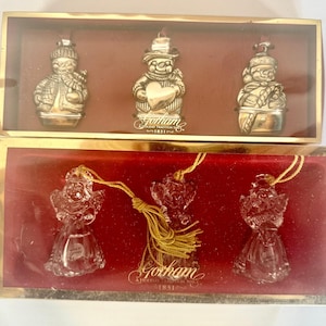 May include: Two boxes of holiday ornaments. The top box contains three silver snowman ornaments. The bottom box contains three clear glass angel ornaments. The boxes have a gold trim and a red interior. The brand name "Gotham" is visible.