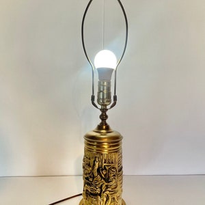 May include: A vintage brass table lamp with a cylindrical base featuring a detailed relief of figures. The lamp has a bulb and a metal shade holder. The lamp is approximately 46 cm tall.