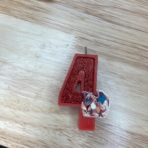 May include: Red glitter birthday candle shaped like the number 4 with a cartoon image of a red and orange Pokemon character on the side.
