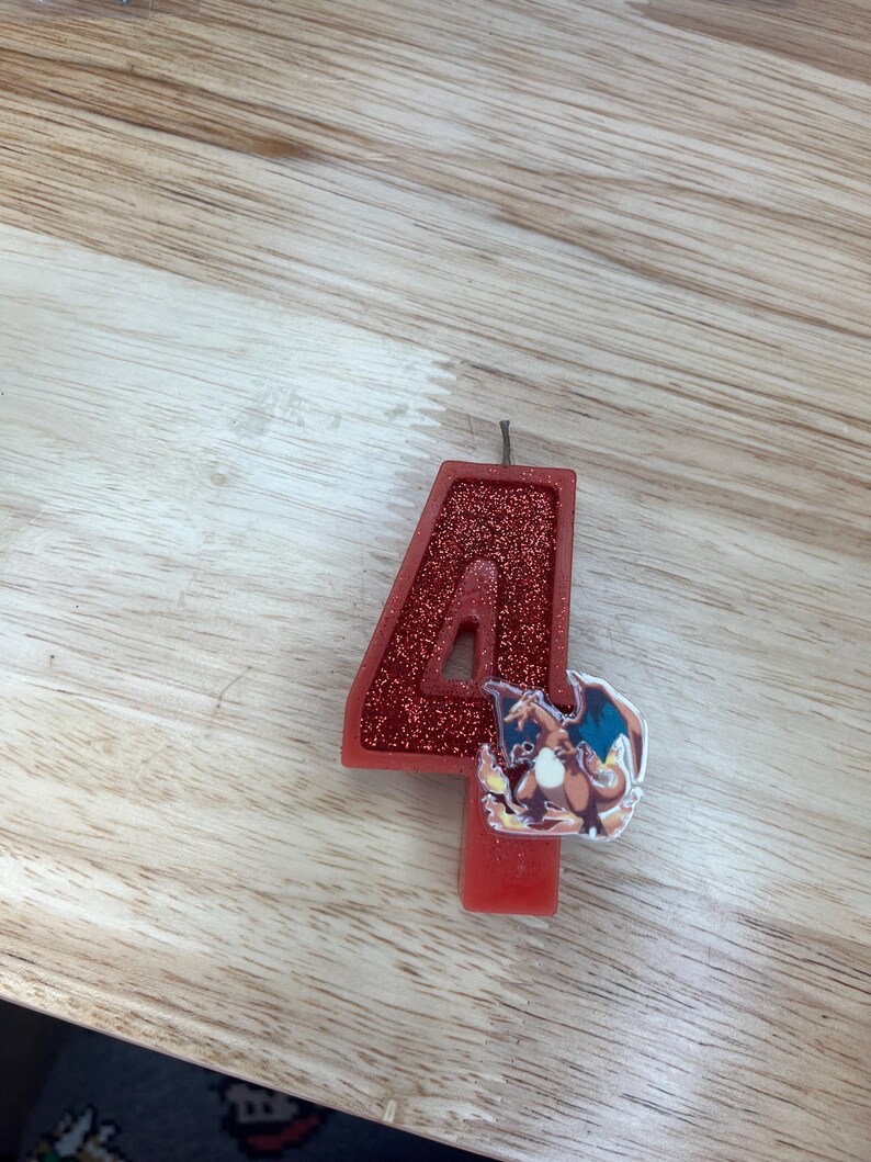 May include: Red glitter birthday candle shaped like the number 4 with a cartoon image of a red and orange Pokemon character on the side.