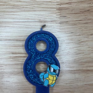 May include: Blue glitter birthday candle shaped like the number eight with a cartoon image of a blue turtle wearing sunglasses.