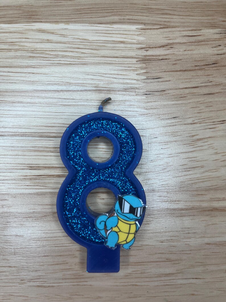 May include: Blue glitter birthday candle shaped like the number eight with a cartoon image of a blue turtle wearing sunglasses.