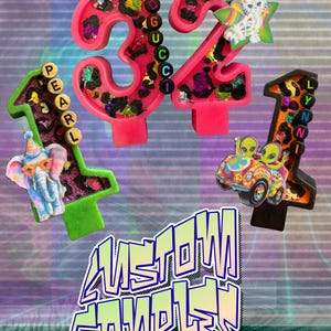 May include: A collection of colorful birthday candles shaped as numbers, including 32, 1, and 1. The candles feature leopard print, glitter, and decorative elements like an elephant, a cat, and aliens in a car. The image also includes the text "AMSTOWN CANDLES".