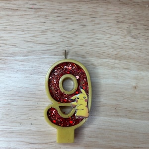 May include: A red and gold glitter birthday candle shaped like the number nine. A cartoon character is on the candle, with a yellow body and red cheeks.