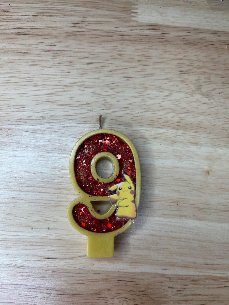 May include: A red and gold glitter birthday candle shaped like the number nine. A cartoon character is on the candle, with a yellow body and red cheeks.