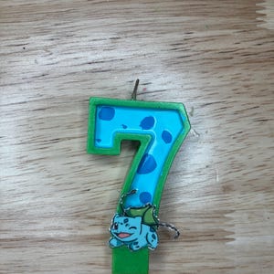 May include: Green and blue birthday candle shaped like the number seven with a cartoon character on the bottom. The character is green and blue and has a white eye.