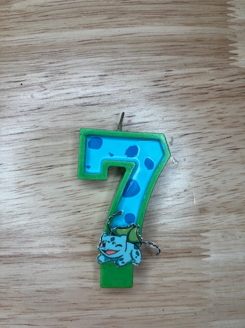 May include: Green and blue birthday candle shaped like the number seven with a cartoon character on the bottom. The character is green and blue and has a white eye.