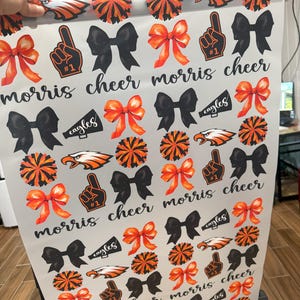 May include: White wrapping paper with a repeating pattern of black bows, orange and black pom-poms, orange and black cheerleaders, and orange and black megaphones with the word "eagles" on them. The text "morris cheer" is repeated throughout the pattern.