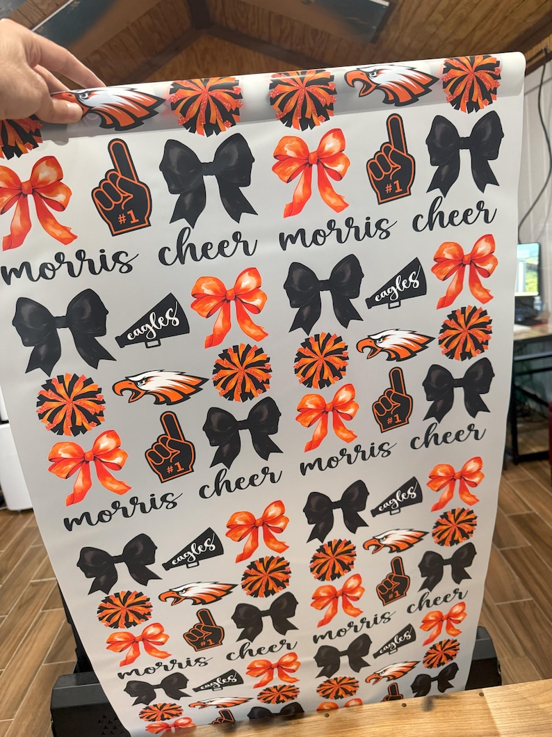 May include: White wrapping paper with a repeating pattern of black bows, orange and black pom-poms, orange and black cheerleaders, and orange and black megaphones with the word "eagles" on them. The text "morris cheer" is repeated throughout the pattern.