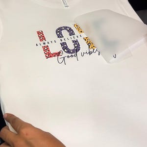 May include: White t-shirt with a colorful "LOVE" graphic featuring a leopard print design. The text "ALWAYS BELIEVE" is written above the graphic and "Good vibes" is written below.