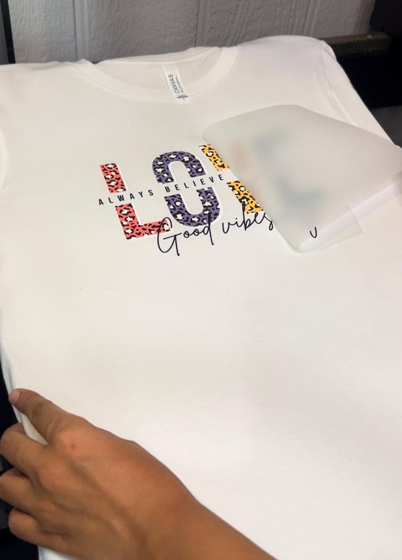 May include: White t-shirt with a colorful "LOVE" graphic featuring a leopard print design. The text "ALWAYS BELIEVE" is written above the graphic and "Good vibes" is written below.