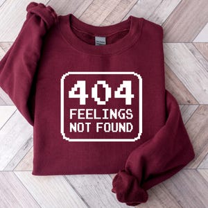 May include: A maroon sweatshirt featuring the text "404 FEELINGS NOT FOUND" in a white, pixelated font. The garment is folded, with the sleeves visible. The design is centered on the chest.
