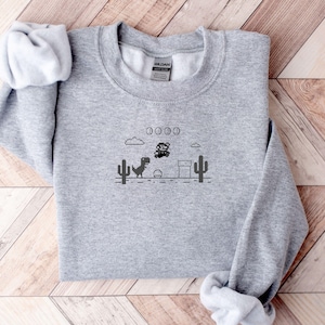 May include: A heather gray sweatshirt featuring a black graphic of a dinosaur and a bear in a video game scene with cacti, coins, and a pipe. The sweatshirt has long sleeves and a crew neck.