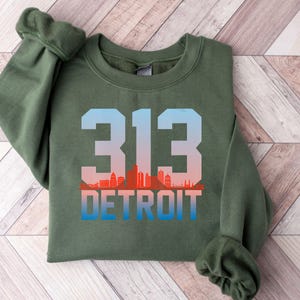 May include: An olive green sweatshirt featuring the number "313" in a gradient blue and red color scheme, with the word "DETROIT" below. The design includes a skyline silhouette. The sweatshirt is made of a soft material.