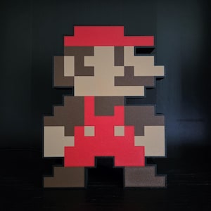 May include: A pixelated Mario figure, featuring a red cap, brown hair, and a tan face. He wears a red jumpsuit with brown shoes. The figure is a 3D rendering of the classic video game character, standing against a black background.