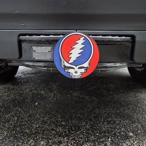 May include: A circular hitch cover featuring the Grateful Dead Steal Your Face logo with a white lightning bolt centered on a blue and red background, attached to the rear bumper of a vehicle above a black towing information sticker.