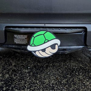 May include: A circular hitch cover featuring a graphic of a green turtle shell with a white rim and a beige underside. The hitch cover is mounted on a black vehicle trailer hitch receiver that displays a black warning label with white text.