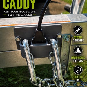 May include: A gray trailer plug caddy mounted to a metal trailer frame, holding a black electrical plug. The caddy features text reading TRAILER PLUG CADDY, KEEP YOUR PLUG SECURE & OFF THE GROUND, STRONG & DURABLE, DRAIN HOLES FOR PLUG, and BUILT TO LAST.