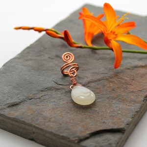 May include: A white teardrop-shaped stone pendant hangs from a copper wire wrapped around a copper spiral. The pendant is on a slate-colored surface with orange flowers in the background.