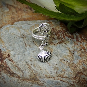May include: A silver ring with a spiral design and a dangling seashell charm. The ring is on a textured brown rock.