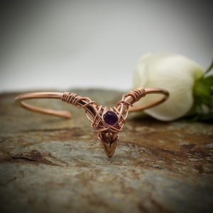 May include: A copper-colored wire-wrapped tiara with a central purple gemstone. The tiara has a V-shape design with intricate wire detailing and is set against a blurred background with a white rose.