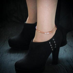 Puede incluir: Una pulsera de tobillo de color cobre se usa sobre una bota negra de tacón alto. La bota tiene una suela de plataforma y un detalle de cordones en el lateral. El fondo es oscuro.