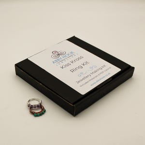 May include: A black square box labeled Abby Hook Jewellery Kiss Kross Ring Kit with a white sticker containing text and two wire-wrapped rings, one featuring purple beads and the other featuring green beads, placed in front of the box.