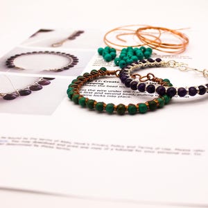 May include: Two handmade beaded bracelets resting on a printed tutorial sheet. One bracelet features green beads wrapped in copper wire, while the other has purple beads wrapped in silver-toned wire. Loose green beads and copper wire are also visible.