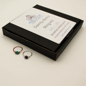May include: A black cardboard box labelled Abby Hook Dainty Boho Ring Kit Jewellery Making Kit with two rings in front. One ring is copper-toned with a turquoise bead, and the other is silver-toned with a purple bead, both featuring wire-wrapped details.