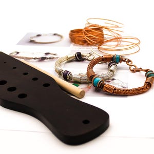May include: A black wooden wire-weaving tool with multiple circular holes, a wooden dowel, two handcrafted wire bracelets featuring purple and turquoise beads, and several coils of copper wire arranged for jewelry making projects.