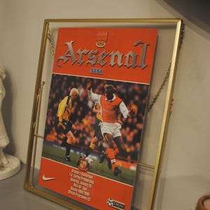 Puede incluir: Un programa de fútbol del Arsenal enmarcado de la temporada 1999/2000. El programa presenta una foto de un jugador con una camiseta roja y blanca, con el texto "Arsenal" y "SEGA" impreso en la portada.