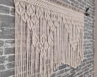 May include: A white macrame wall hanging with a geometric pattern. The hanging is made of cotton cord and has a fringe at the bottom.