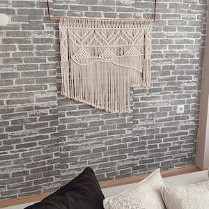 May include: A white macrame wall hanging with a geometric pattern. The hanging is made of cotton cord and is attached to a wooden dowel.
