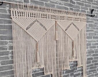 May include: A beige macrame wall hanging with a geometric pattern and fringe. The hanging is made of natural fibres and has a rustic, bohemian style.