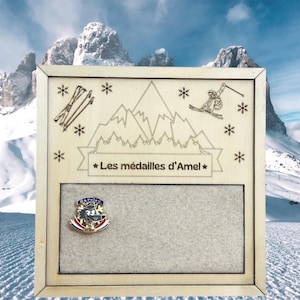 Peut inclure: Un panneau d'affichage carré en bois avec des motifs gravés au laser représentant des montagnes, des skis et des flocons de neige. Une bannière centrale porte le texte "Les médailles d'Amel" au-dessus d'un panneau en feutre gris avec une épinglette.