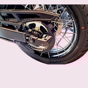 May include: Close-up of a black motorcycle chain guard with a silver chain and sprocket. The guard has a black plastic cover with a silver logo that says "Snolade".