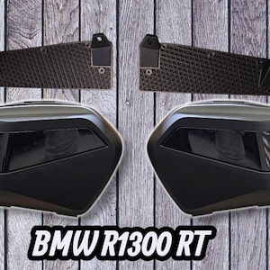 Pode incluir: Duas malas laterais pretas para motocicletas e suportes de montagem são exibidos em um fundo de grão de madeira. As malas têm um design elegante e angular com acabamento preto brilhante. O texto "BMW R1300 RT" está na parte inferior.