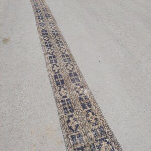 May include: A long, narrow, vintage rug with a repeating geometric pattern in navy blue and beige. The rug is rectangular and appears to be made of woven fabric. The rug is placed on a grey concrete surface.