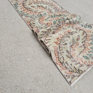 Könnte beinhalten: Ein rechteckiger Läufer mit einem verblassten Blumenmuster in Beige, Orange, Grün und Schwarz. Die Läufer ist am Ende aufgerollt und zeigt seine Textur und sein Design. Der Teppich ist wahrscheinlich aus gewebtem Material.