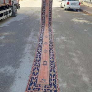 Op de afbeelding: Een lange, smalle vintage loper met een centraal perzikkleurig veld en donkerblauwe randen. De loper heeft geometrische en bloemmotieven, met een rode franje aan het uiteinde. De loper wordt buiten op een betonnen ondergrond getoond.