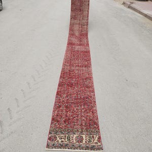 May include: A long, rectangular red rug with an intricate floral pattern. The rug has a faded appearance, with a border of beige, green, and blue floral designs. The rug is rolled out on a gray surface.