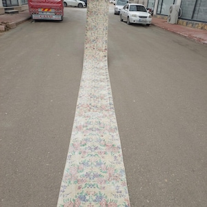 Puede incluir: Una alfombra larga y rectangular con un estampado floral en tonos rosa, azul y verde sobre un fondo crema. La alfombra está desenrollada en una calle, mostrando su longitud y diseño. Los bordes de la alfombra están ligeramente deshilachados.
