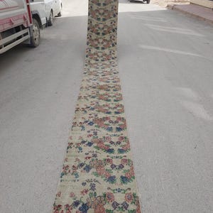 Pode incluir: Um tapete longo e retangular com um padrão floral em tons de rosa, azul e verde sobre um fundo bege. O tapete tem uma borda bordô e é exibido ao ar livre em uma superfície pavimentada.