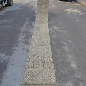 May include: A long, rectangular beige rug with an intricate floral pattern in shades of brown, red, and green. The rug is rolled out on a gray surface, showcasing its detailed design and border.