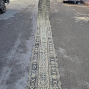 May include: A long, rectangular rug with a detailed, symmetrical pattern. The rug features a central design with floral and geometric motifs in shades of blue, beige, and cream. The rug is displayed outdoors on a gray surface.