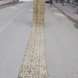 May include: A long, narrow, beige rug with a detailed floral pattern in dark brown. The rug is held up vertically, displaying its length and design. The rug appears to be made of a woven material, suitable for a hallway or entryway.