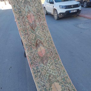 Puede incluir: Una alfombra de pasillo larga y estrecha con un patrón floral y geométrico complejo y repetitivo en tonos apagados de verde salvia, terracota descolorido y marrón oscuro. La alfombra tiene una textura desgastada y envejecida con pelo visible.