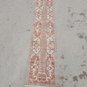 May include: A rectangular runner rug with an intricate pattern. The rug features a central design with repeating geometric shapes in shades of red, cream, and blue. The rug's border is a darker red, and the overall design has a vintage aesthetic.