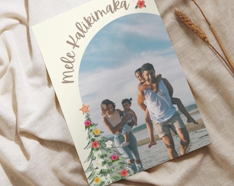 May include: A Christmas card with a tropical theme featuring a family of four standing on a beach. The card has a green and white Christmas tree with pink and yellow flowers. The text "Mele Kalikimaka" is written in brown on the card.