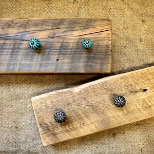 May include: Two rustic wooden coat racks with decorative knobs. One rack has three turquoise knobs, the other has two dark metal knobs. The wood has a weathered, natural finish, and the knobs add a touch of color and detail.